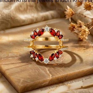 Puede incluir: Un protector de anillo de oro con dos bandas paralelas conectadas por barras verticales. Las secciones superior e inferior están adornadas con granates rojos de talla marquesa y pequeños diamantes blancos redondos en un patrón floral.