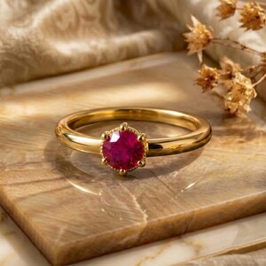 Puede incluir: Un anillo de oro con una piedra preciosa roja oscura, redonda y facetada, montada en un engaste de seis garras. La banda es lisa y pulida, con la piedra central sujeta por pequeñas garras redondeadas que crean un borde texturizado.
