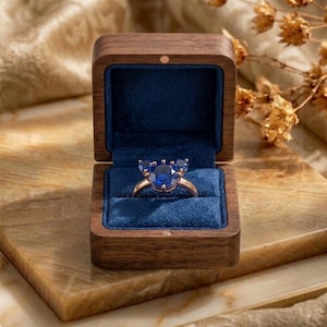 May include: A ring with a central round blue gemstone flanked by two smaller round blue gemstones, set on a metallic band. The ring is displayed inside a square wooden box with a dark blue velvet interior lining and a hinged lid.