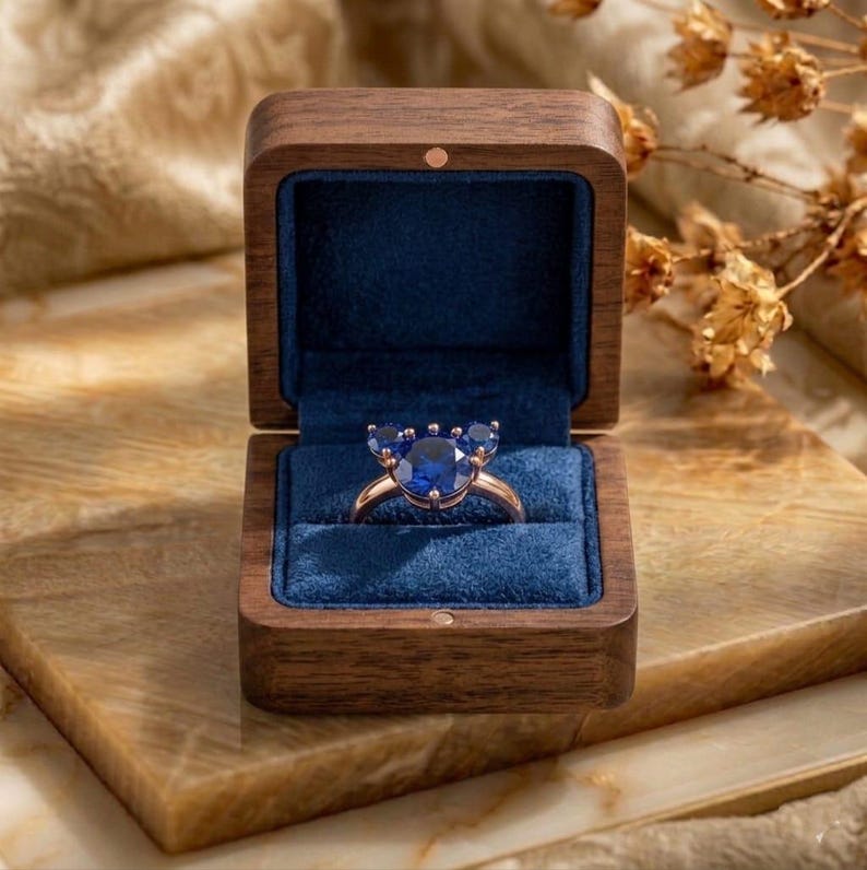 May include: A ring with a central round blue gemstone flanked by two smaller round blue gemstones, set on a metallic band. The ring is displayed inside a square wooden box with a dark blue velvet interior lining and a hinged lid.