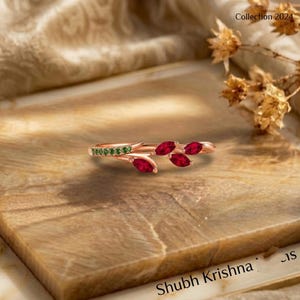 May include: A rose gold ring featuring a band adorned with small green stones and three marquise-cut red stones arranged in a leaf-like pattern. The ring has a delicate, open-ended design that curves around the finger with a polished metallic finish.