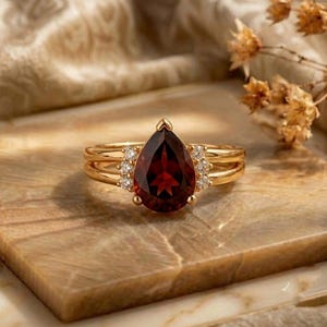 Può includere: Un anello in oro con una grande pietra preziosa granato rosso scuro a forma di pera come elemento centrale. La fascia è realizzata in oro lucido e include tre piccole pietre d'accento rotonde e trasparenti su ogni lato della montatura.