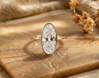 May include: A gold ring featuring a large, oval-cut, clear gemstone set in a smooth bezel setting. The band is thin and polished, showcasing a reflective surface. The design is centered, highlighting the elongated shape of the central stone.