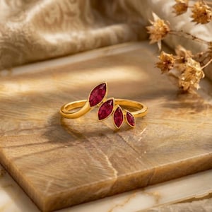 May include: A gold ring featuring four marquise-cut red gemstones arranged in a cluster along the top of the band. The stones are set in a row, decreasing in size from left to right, creating a curved, leaf-like shape on the polished gold band.