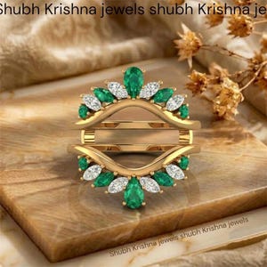 May include: A gold ring guard featuring two curved bands connected by vertical bars. The design is adorned with alternating pear-cut green stones and marquise-cut clear stones arranged in a symmetrical, fan-like pattern along the outer edges.