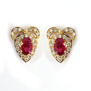 May include: A pair of gold heart-shaped earrings with a red ruby gemstone in the center of each earring. The ruby is surrounded by a halo of smaller diamonds.
