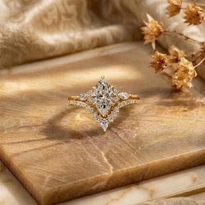 Pode incluir: Um anel de ouro com uma pedra preciosa central transparente de corte marquise rodeada por um halo de pedras redondas mais pequenas. A banda tem forma de V e está incrustada com pequenas pedras transparentes adicionais, criando um design simétrico.