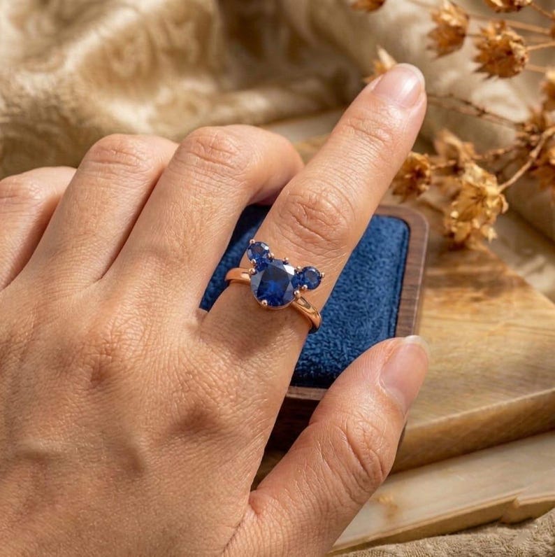 May include: A rose gold ring featuring a central oval-cut blue sapphire flanked by two smaller round-cut blue sapphires, arranged in a three-stone cluster design that resembles a mouse head silhouette on a thin polished metal band.