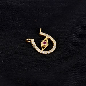 May include: Gold horseshoe pendant with a diamond-encrusted design and a red gemstone in the center, resembling an eye.