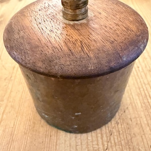 May include: A cylindrical container with a hammered copper body and a circular wooden lid. The lid features a small, central metallic knob with a tiered, rounded design. The copper surface shows a weathered, mottled patina with visible texture.