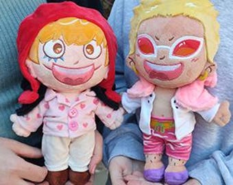 May include: Two plush dolls of anime characters are held by a person. The doll on the left has orange hair, a red hood, and a pink pajama top with heart designs. The doll on the right has blonde hair, pink sunglasses, and a white jacket.