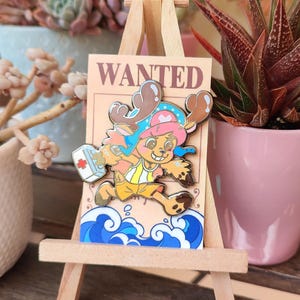 Puede incluir: Un colorido pin de esmalte con un personaje de dibujos animados con cuernos marrones, un sombrero azul y pantalones cortos amarillos. El pin está sobre un caballete de madera con un fondo de póster de "SE BUSCA". El personaje corre sobre olas azules.