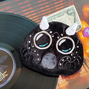 Puede incluir: Un juguete de peluche negro con cuernos blancos y ojos grandes, sobre un disco de vinilo. También se ven una púa morada y un billete de dólar. El disco tiene una etiqueta con el logo de la banda.