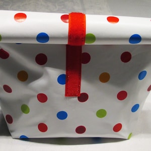 Könnte beinhalten: Weiße Lunch-Tasche mit einem bunten Polka-Dot-Muster in Rot, Blau, Grün und Gelb. Die Tasche wird mit einem roten Riemen gesichert. Die Tasche besteht aus einem flexiblen Material.