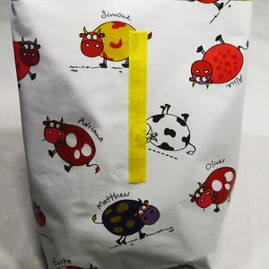 Könnte beinhalten: Weißer Stoffbeutel mit gelbem Besatz und einem gelben vertikalen Streifen. Die Tasche ist mit Cartoon-Kühen in Rot, Gelb, Schwarz und Lila verziert, jede mit einem Namen. Zu den Namen gehören Simone, Alice, Adriana, Oliver, Matthew und Evita.