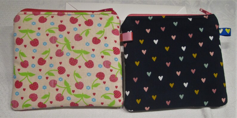 以下が含まれることがあります： ジッパー付きポーチが2つ。1つはベージュの背景にチェリー柄、もう1つは濃い青の背景にハート柄です。