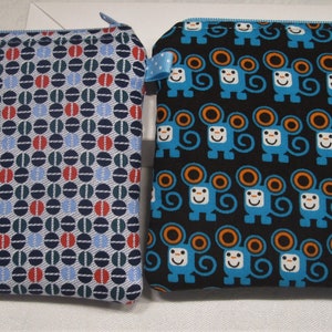 以下が含まれることがあります： 異なるパターンを持つジッパー付きポーチ2つ。1つのポーチは青、赤、緑の幾何学模様。もう1つのポーチは黒地にオレンジ色の耳を持つ青い猿の繰り返しパターン