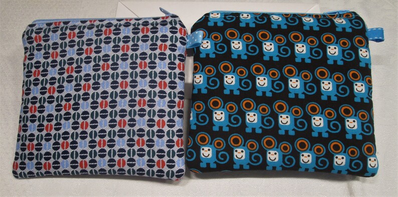以下が含まれることがあります： 異なるパターンを持つジッパー付きポーチ2つ。1つのポーチは青、赤、緑の幾何学模様。もう1つのポーチは黒地にオレンジ色の耳を持つ青い猿の繰り返しパターン