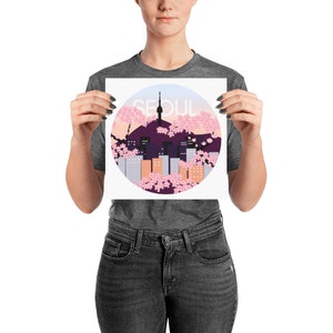 May include: A white print with a circle design featuring a cityscape of Seoul, South Korea, with a pink and purple cherry blossom background. The text "SEOUL" is written in white letters on the print.