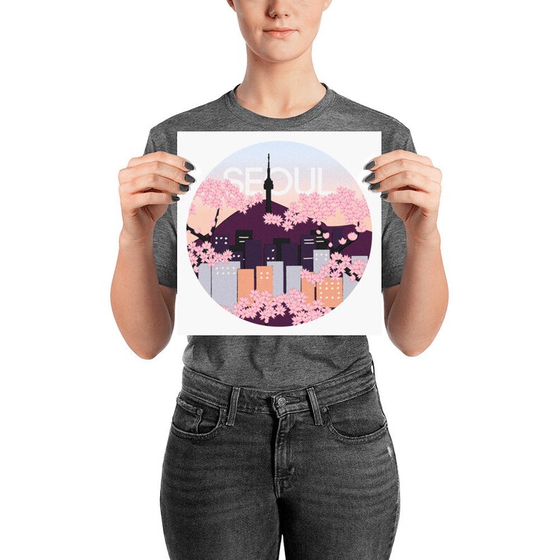 May include: A white print with a circle design featuring a cityscape of Seoul, South Korea, with a pink and purple cherry blossom background. The text "SEOUL" is written in white letters on the print.