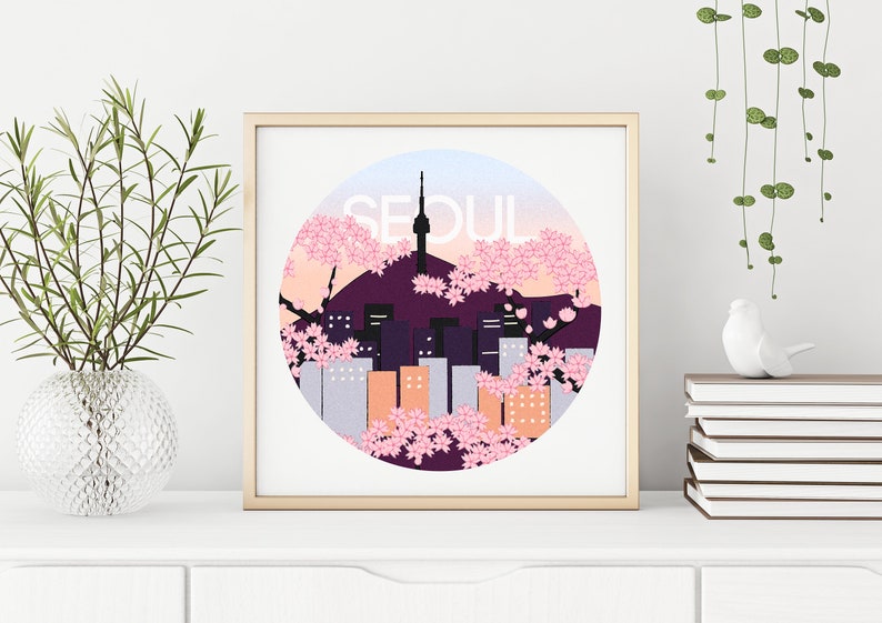 May include: A framed print with a circular design featuring a cityscape of Seoul, South Korea, with a tower and cherry blossoms in pink and purple hues. The word "SEOUL" is written in white letters on a light blue background.
