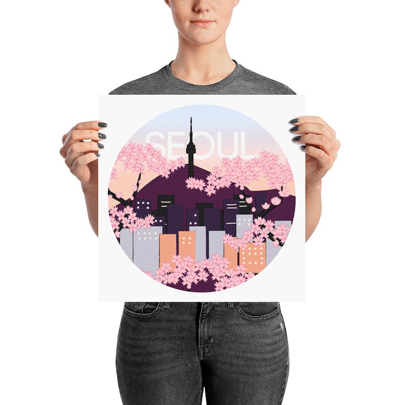May include: A white poster with a circle in the center. The circle has a pink and purple cityscape with a tower in the center. The text "SEOUL" is above the cityscape in white letters.