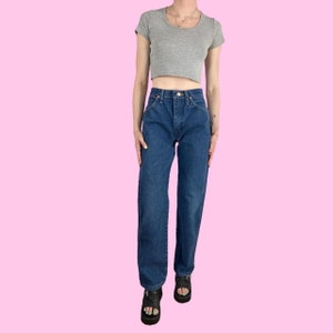 Puede incluir: Un par de jeans azules de tiro alto y pierna recta con una camiseta gris claro recortada. La modelo lleva sandalias negras.