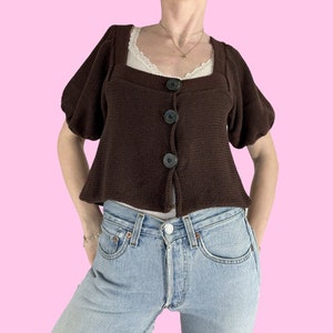 May include: A brown knitted cardigan with puffed sleeves and a square neckline. The cardigan has four black buttons down the front.