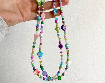 Puede incluir: Dos collares de cuentas superpuestos con cuentas y dijes coloridos, incluyendo estrellas, corazones, caritas sonrientes y un dado. Los collares están hechos con cuentas verdes, azules, rosas, moradas y blancas.