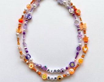 May include: A layered beaded necklace with orange, white, and purple beads. The necklace features various charms, including butterflies, flowers, smiley faces, and a yin-yang symbol. The necklace also includes the words "FIDO" and "SIR" spelled out in white beads.