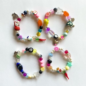 May include: Four colorful beaded bracelets with various charms, including a butterfly, a cat, a lemon, a turtle, a smiley face, a chili pepper, an eye, a heart, a 8-ball, a star, a slice of lemon, and a cherry.