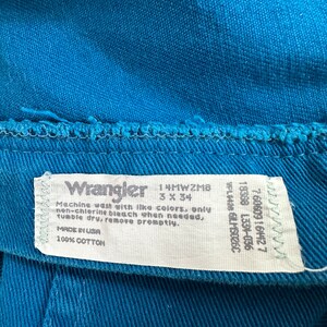 Może przedstawiać: Biała metka pielęgnacyjna Wrangler z niebieskim tekstem. Na metce widnieje napis "Machine wash with like colors, only non-chlorine bleach when needed, tumble dry, remove promptly. Made in USA 100% Cotton". Metka zawiera r&oacute;wnież rozmiar 3x34 i kod produktu 14MWZMB.