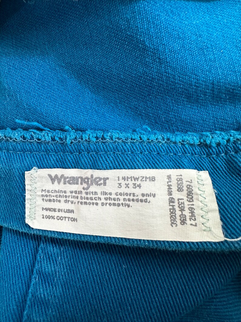 Może przedstawiać: Biała metka pielęgnacyjna Wrangler z niebieskim tekstem. Na metce widnieje napis "Machine wash with like colors, only non-chlorine bleach when needed, tumble dry, remove promptly. Made in USA 100% Cotton". Metka zawiera r&oacute;wnież rozmiar 3x34 i kod produktu 14MWZMB.