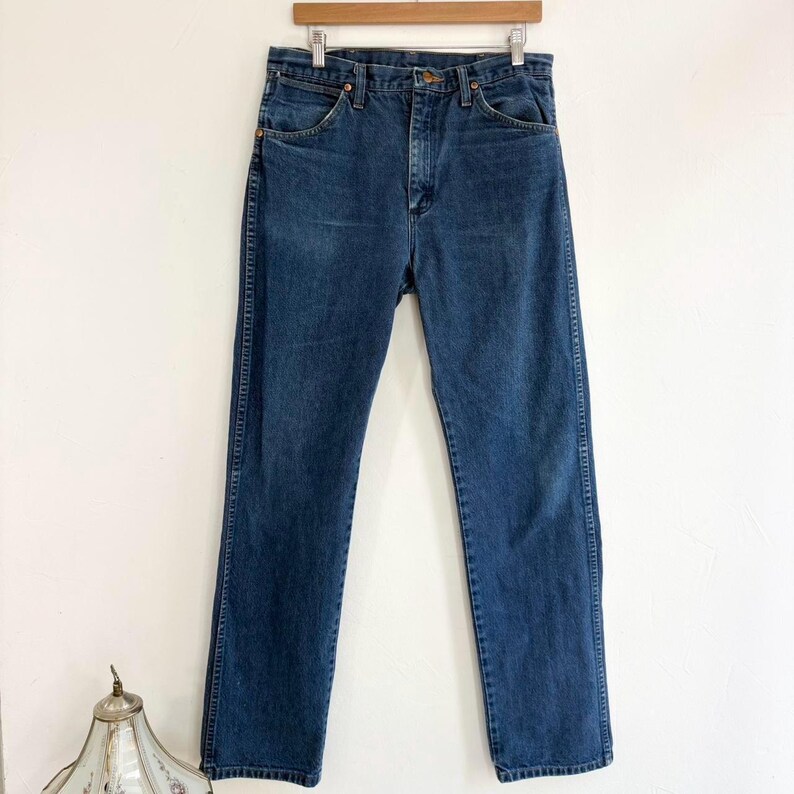 May include: Dark wash denim jeans hanging on a wooden hanger. The jeans have a classic five-pocket design, a button and zipper closure, and a straight leg. The denim is a medium weight, with subtle fading and wear.