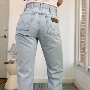 Op de afbeelding: Lichtblauwe denim jeans met een hoge taille en een klassiek ontwerp met vijf zakken. De jeans hebben het Wrangler-logo op de achterzak en een bruine leren patch. De lichte wassing en de rechte pijpen geven een vintage esthetiek.
