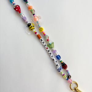 May include: A beaded lanyard featuring a variety of colourful charms including a strawberry, a bee, a green butterfly, a ladybug, and smiley faces. The beads spell out MRS BROOKE with a red heart between the words, ending in a gold metal clasp.
