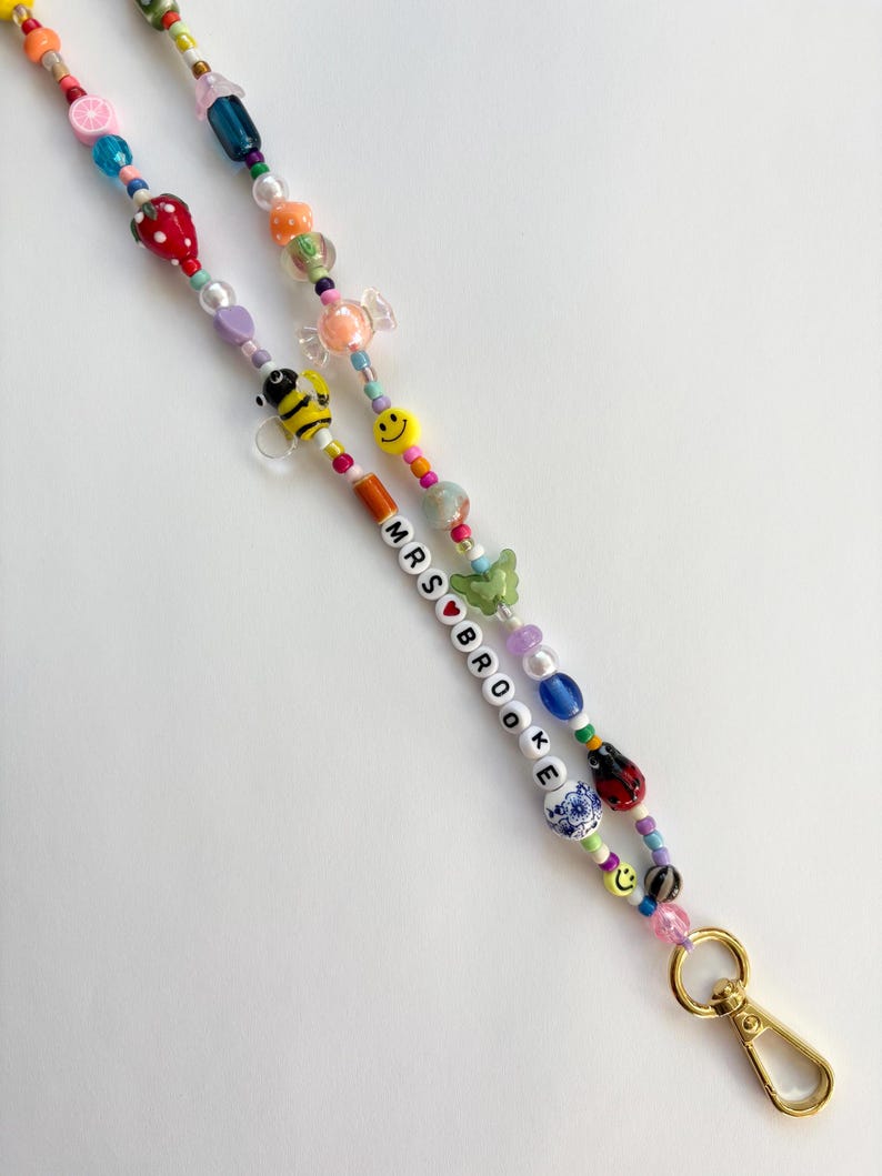 May include: A beaded lanyard featuring a variety of colourful charms including a strawberry, a bee, a green butterfly, a ladybug, and smiley faces. The beads spell out MRS BROOKE with a red heart between the words, ending in a gold metal clasp.
