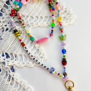 May include: A colourful beaded lanyard featuring a variety of shapes, including smiley faces, a bee, a ladybug, and fruit charms. The beads are strung on a cord with a pink breakaway clasp and a gold-tone metal lobster clasp. The name MRS BROOKE is spelled out in white letter beads with a red heart.