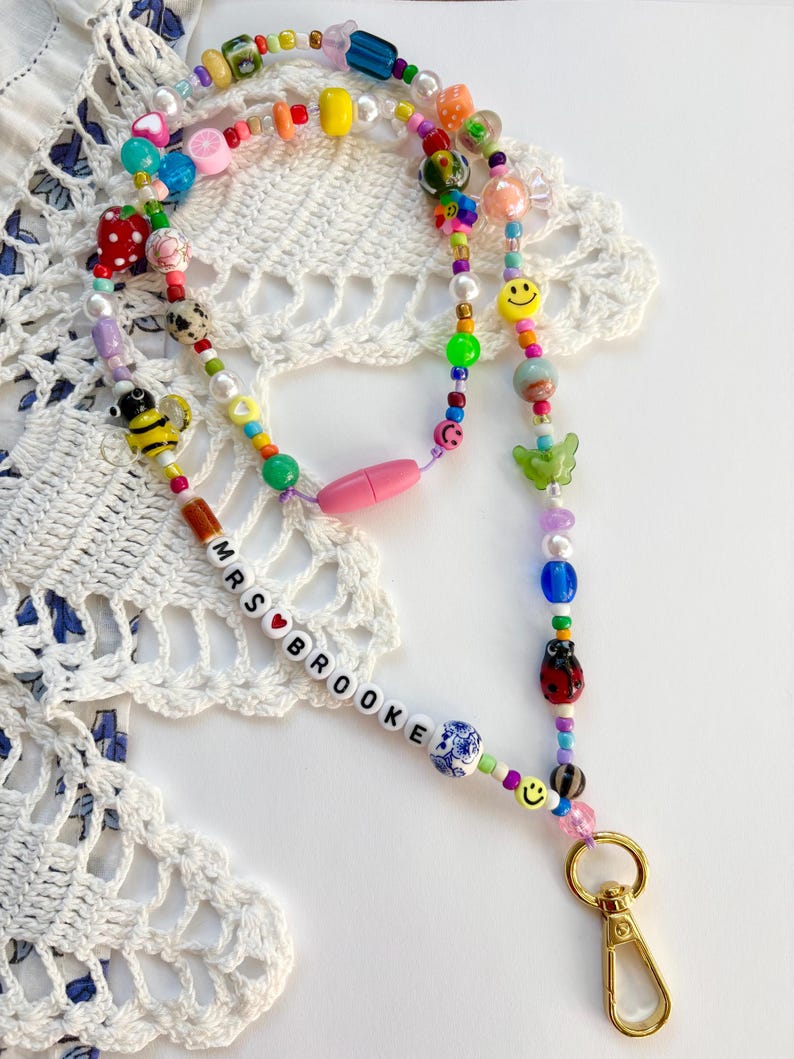 May include: A colourful beaded lanyard featuring a variety of shapes, including smiley faces, a bee, a ladybug, and fruit charms. The beads are strung on a cord with a pink breakaway clasp and a gold-tone metal lobster clasp. The name MRS BROOKE is spelled out in white letter beads with a red heart.