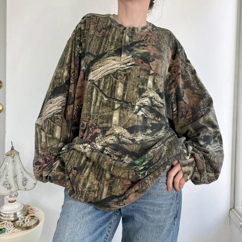 K&ouml;nnte beinhalten: Lang&auml;rmeliges Camouflage-Hemd mit Knopfleiste und Brusttasche. Das Hemd hat ein Muster aus braunen, gr&uuml;nen und beigen Farbt&ouml;nen, das einer Waldszene &auml;hnelt. Das Hemd wird mit einer blauen Jeans kombiniert.