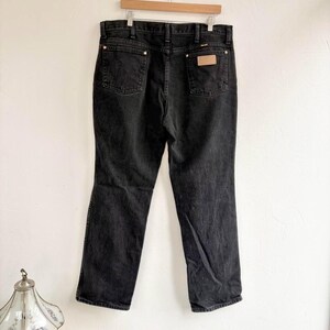 May include: Black denim jeans hanging on a wooden hanger. The jeans feature two back pockets, a small leather patch, and copper-coloured rivets. The jeans are a classic, straight-leg style.