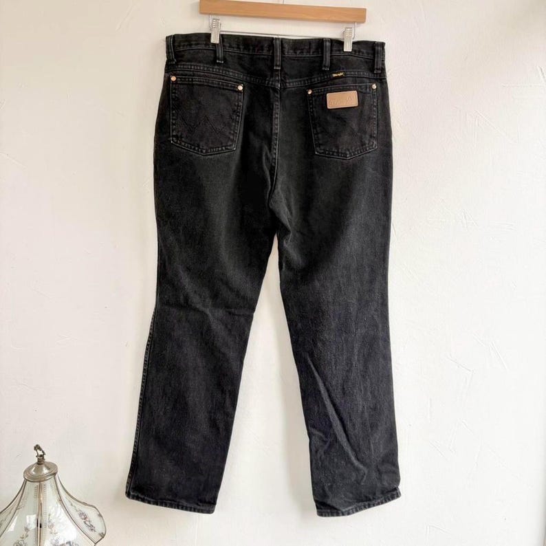 May include: Black denim jeans hanging on a wooden hanger. The jeans feature two back pockets, a small leather patch, and copper-coloured rivets. The jeans are a classic, straight-leg style.