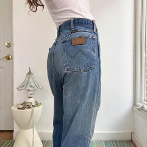 May include: Light blue denim Wrangler jeans with a brown leather patch on the back pocket. The jeans have a classic five-pocket design and a high waist. The jeans are worn and faded, giving them a vintage look.