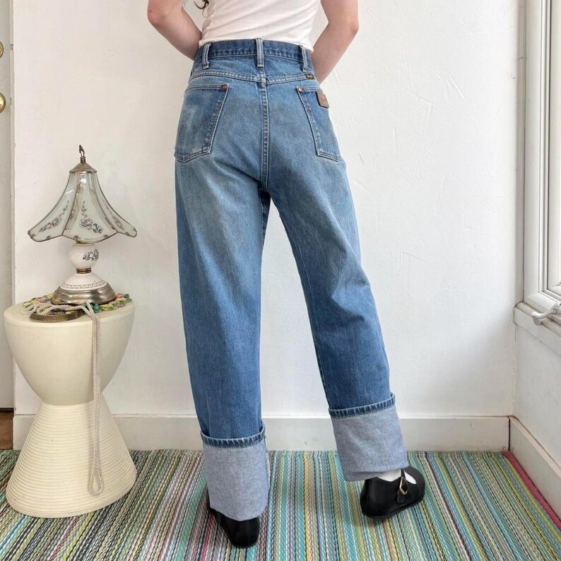 May include: Light blue denim jeans with a cuffed hem. The jeans have a classic five-pocket design and a brown leather patch on the back. The model is wearing black shoes. The jeans are a vintage style.