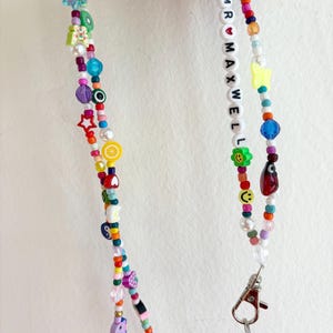 May include: A colorful beaded lanyard with a silver clasp. The beads are in various shapes and sizes, including stars, hearts, flowers, and smiley faces. The lanyard also features white beads with the letters "MR MAXWELL" spelled out.