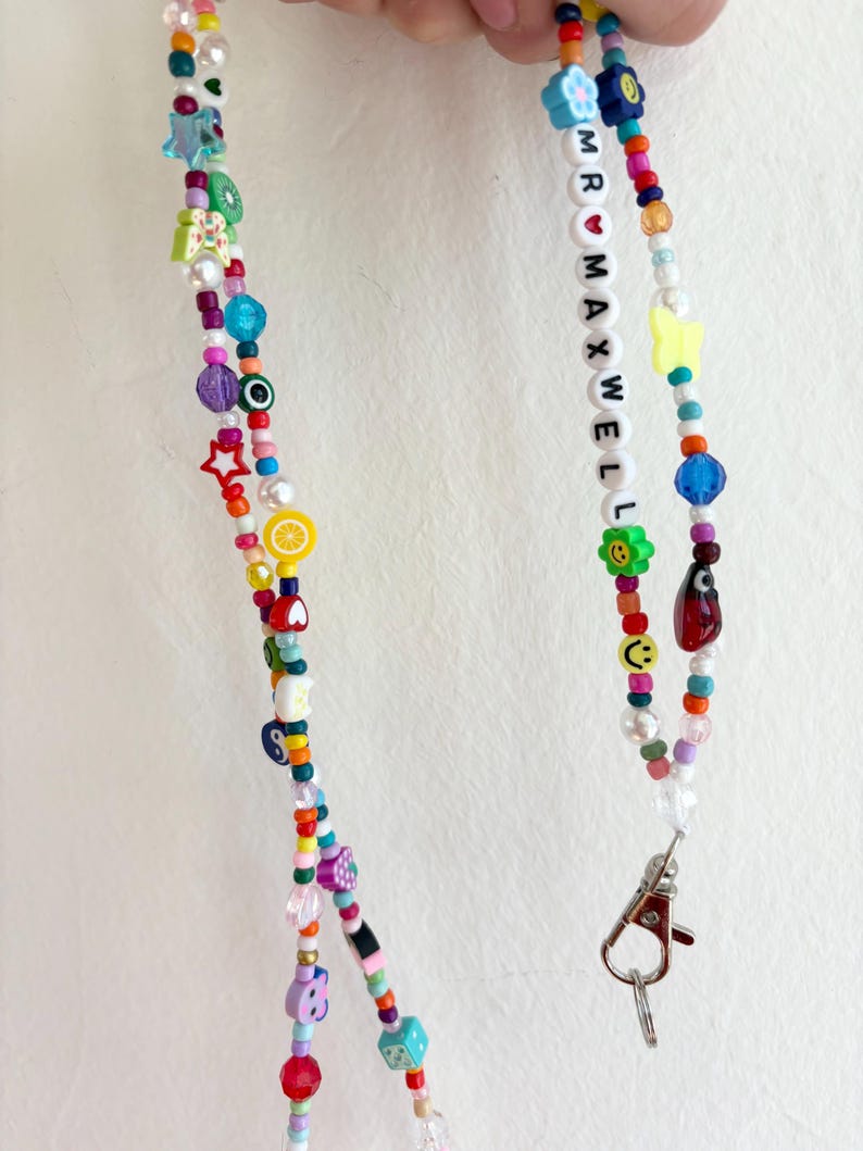 May include: A colorful beaded lanyard with a silver clasp. The beads are in various shapes and sizes, including stars, hearts, flowers, and smiley faces. The lanyard also features white beads with the letters "MR MAXWELL" spelled out.