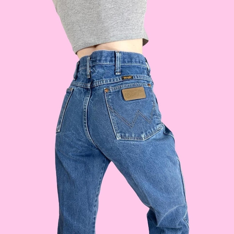 May include: Blue denim Wrangler jeans with a high waist and a brown leather patch on the back pocket. The jeans have a classic five-pocket design and a straight leg. The Wrangler logo is visible on the patch.
