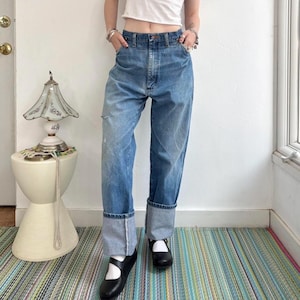 May include: Light blue denim jeans with a relaxed fit and cuffed hems. The jeans have a classic five-pocket design and a faded wash. The model is wearing black Mary Jane shoes. The jeans are a vintage style.