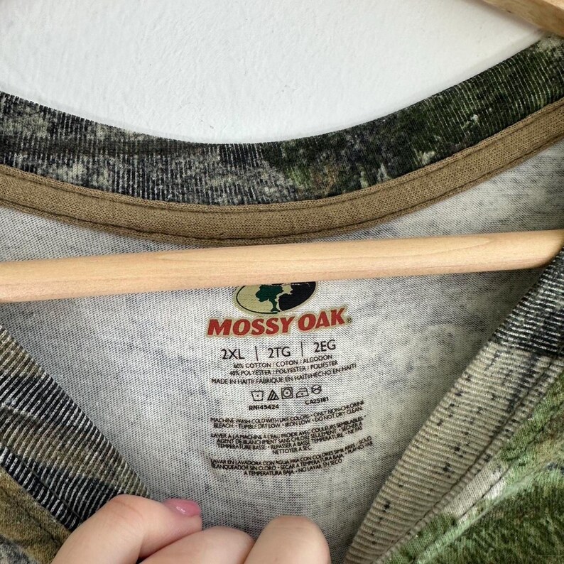 May include: A Mossy Oak brand long-sleeve shirt in a camouflage pattern with a white base. The shirt has a brown trim around the neck and a tag with the brand name, size, and fabric content. The tag reads "Mossy Oak", "2XL", "2TG", "2EG", "40% Cotton", "40% Polyester", "Made in Haiti", "Fabriqu&eacute; en Ha&iuml;ti", "Hecho en Hait&iacute;", "RN145424", "CA52519".