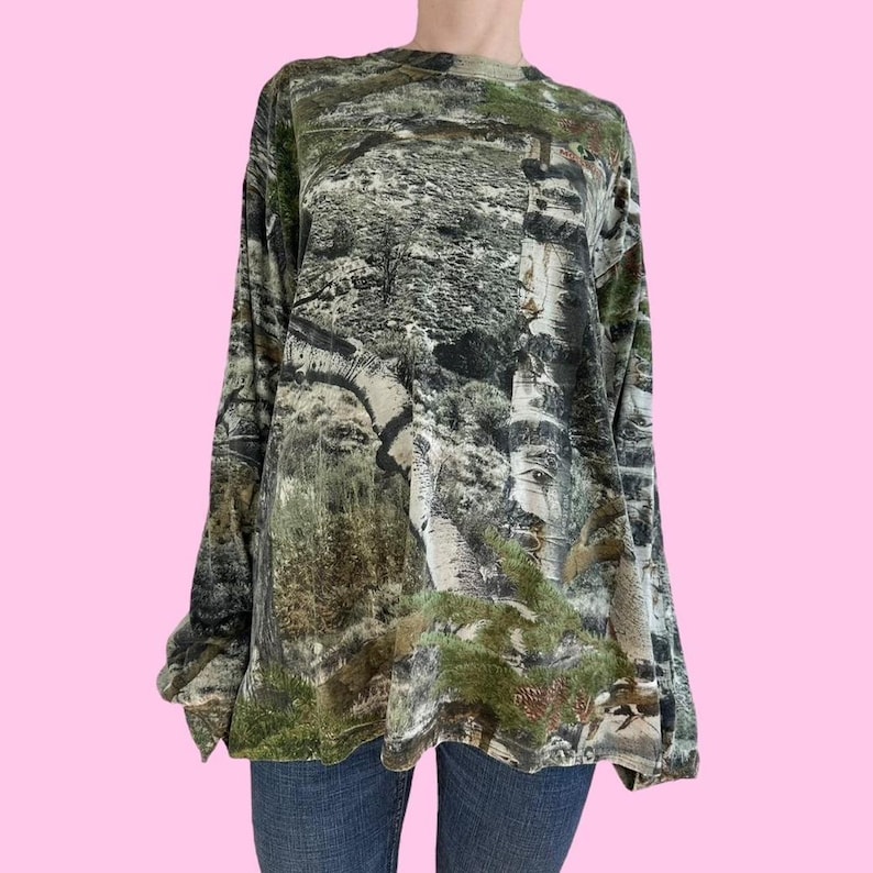 May include: A long-sleeved shirt with a camouflage pattern in shades of green, brown, and white. The pattern features a variety of trees and foliage.